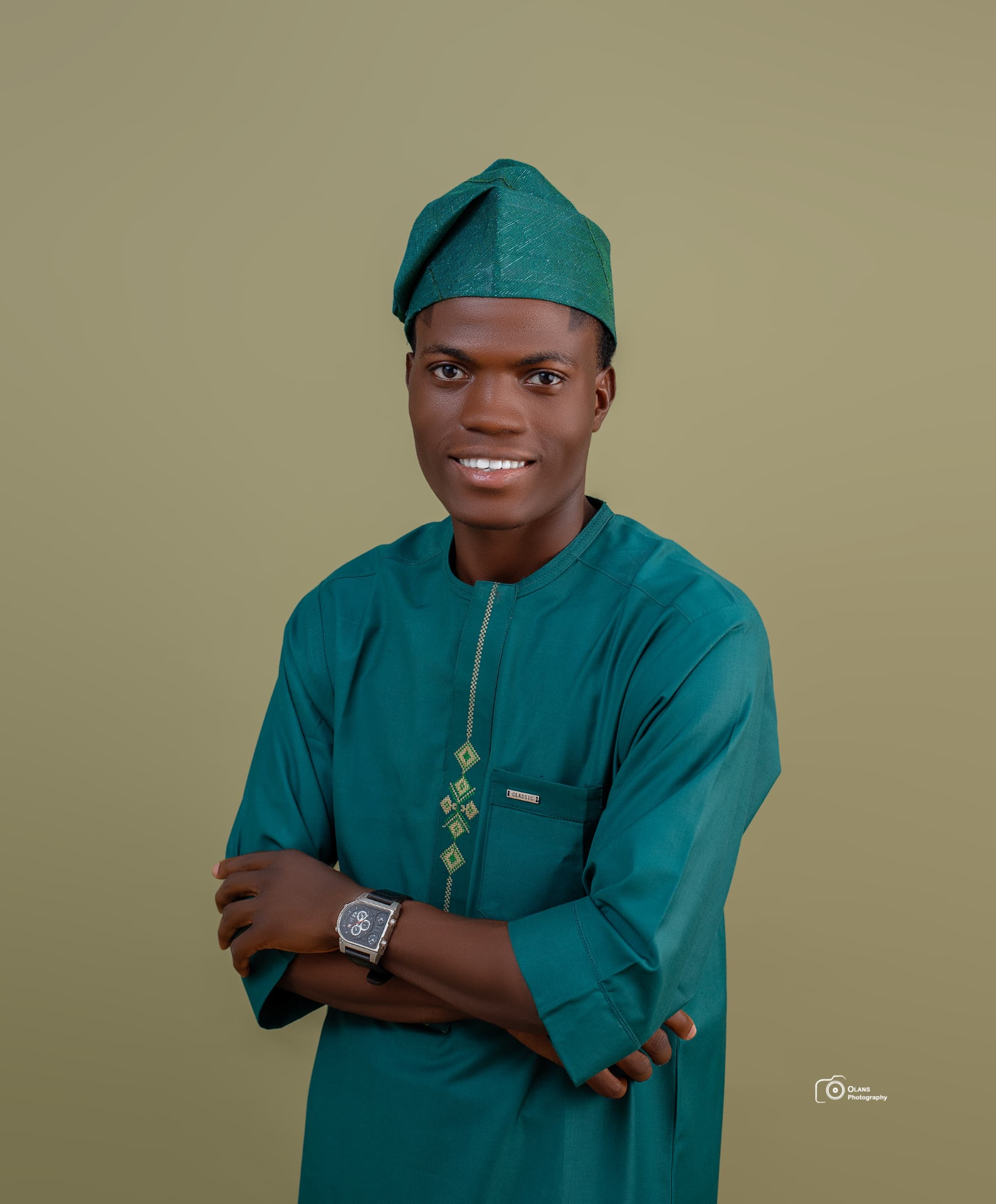 OGM smiling in green traditional attire and fila cap, arms folded